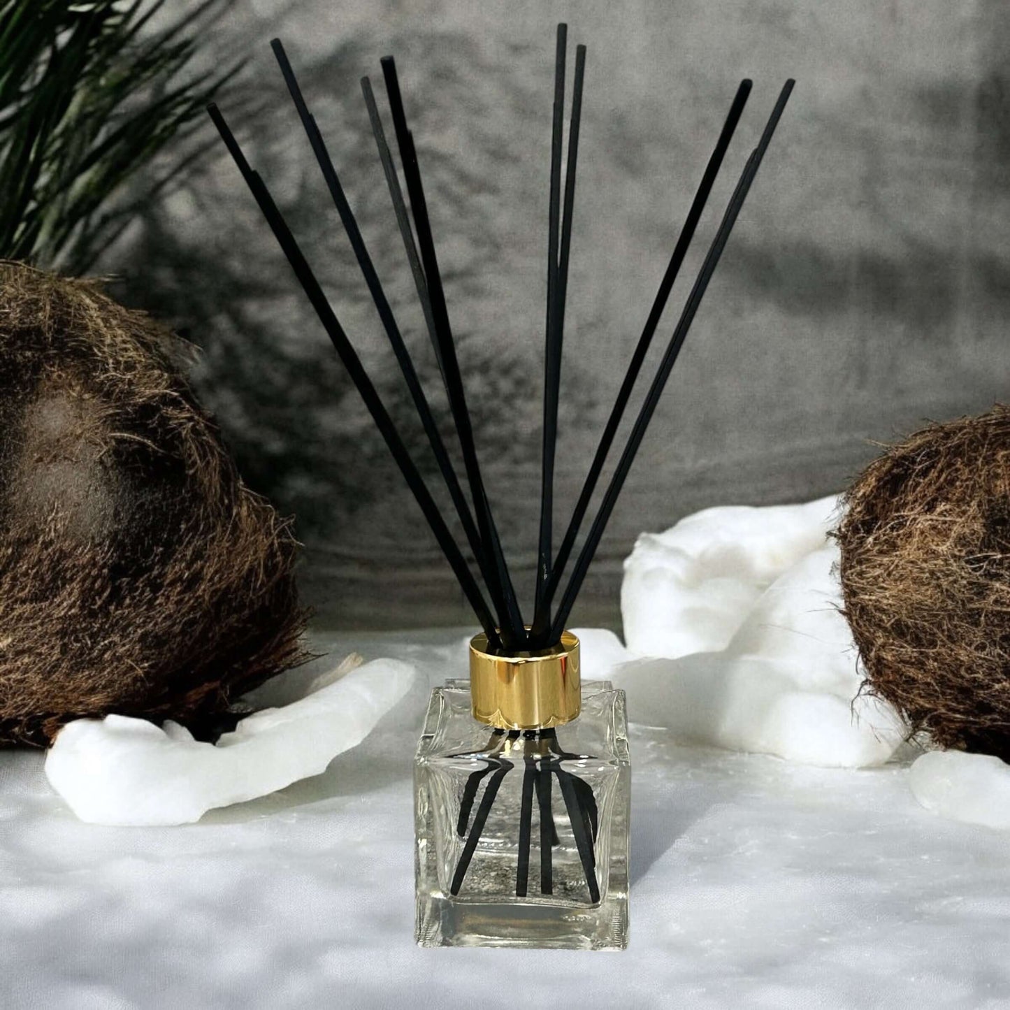 Handmade reed diffuser with black sticks in a glass bottle, surrounded by coconut shells and soft white decoration.