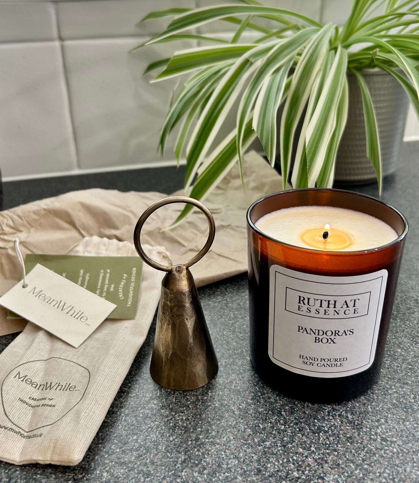 Handmade brass candle snuffer beside lit Ruth at Essence soy candle, styled with natural textures and a houseplant for an eco-friendly home aesthetic.